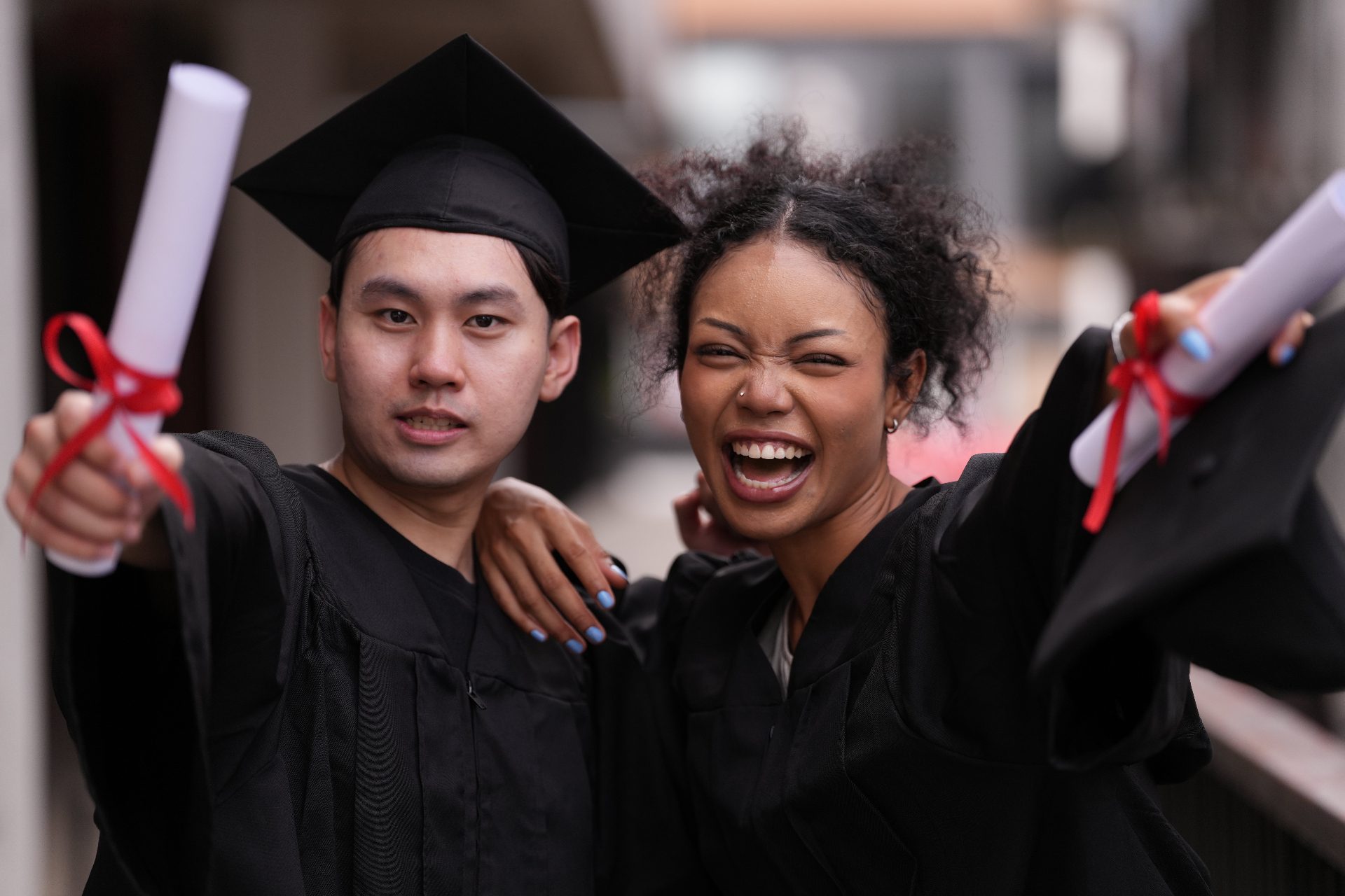 Two Class of 2026 graduates in caps and gowns, arms raised, holding diplomas tied with red ribbon on commencement day