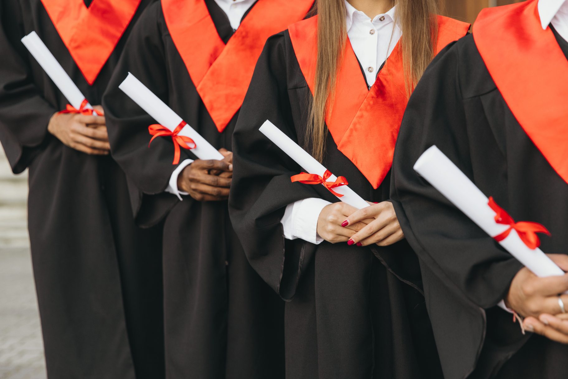 A row of graduates in black robes with red sashes, holding diplomas tied with red ribbons